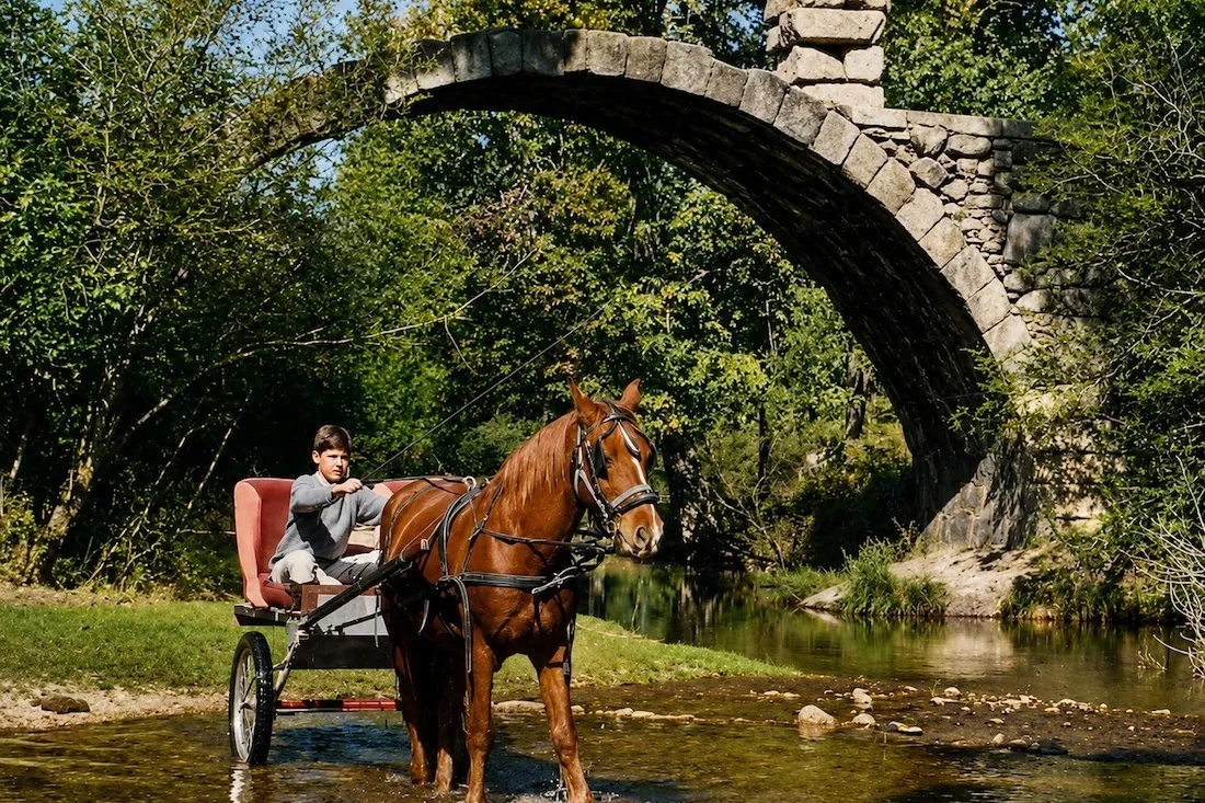 Primer prototipo del Coche de Caballos Navarro cruzando un río bajo un puente de piedra, conducido por un niño